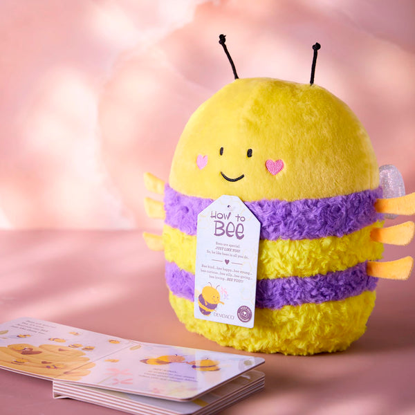 A yellow and purple striped plush bee toy with orange legs and antennae, featuring a hangtag that reads 'How to Bee', set against a pink background with an open board book in front.